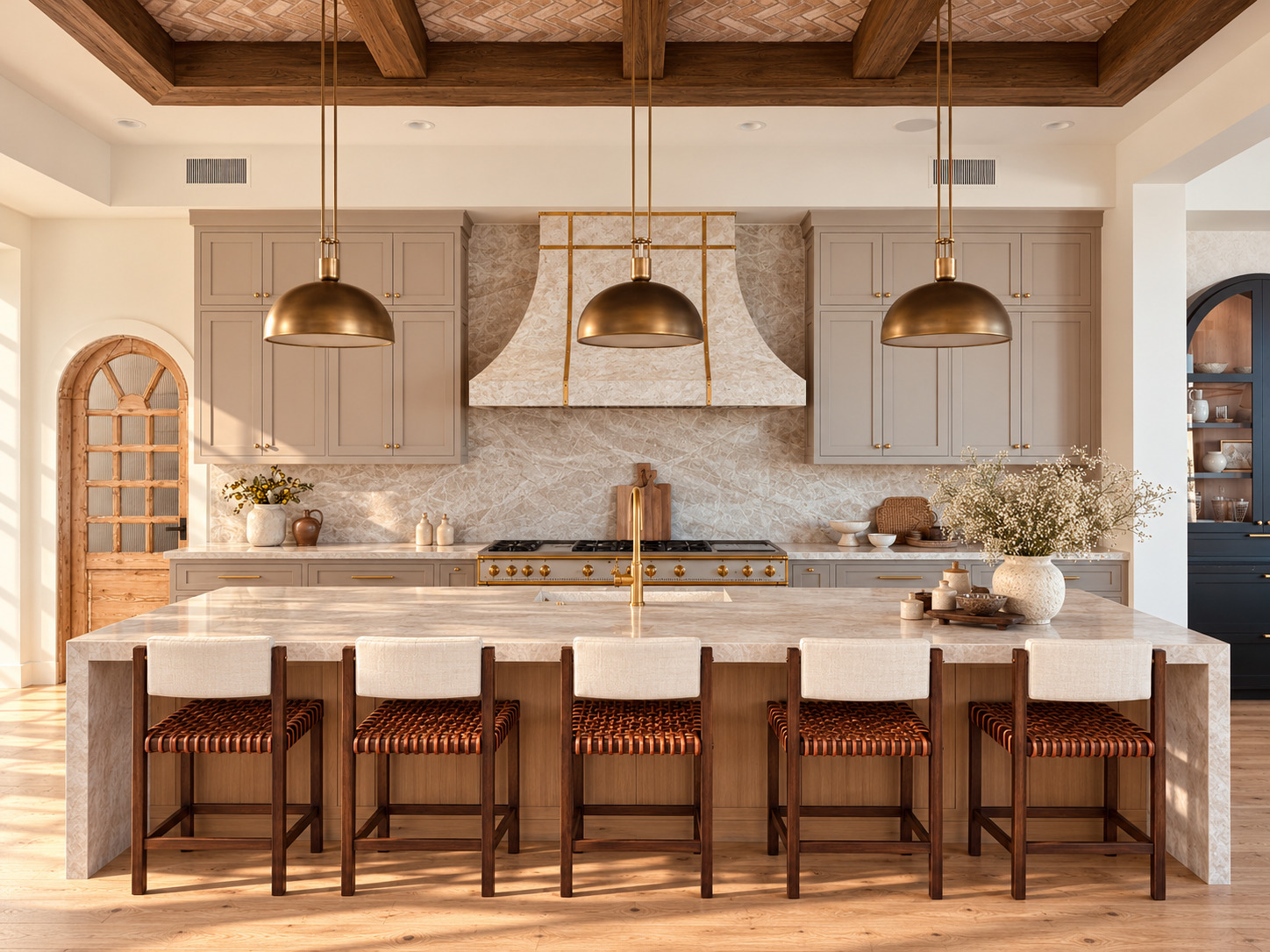 A head-on kitchen view with a slim stone waterfall island, five woven leather stools, and three unlit brass dome pendants on paired rods. Dark timber frames a herringbone brick ceiling. An antique timber pantry door with weathered wood and textured glass sits left of a French range beneath a curved stone hood with brass trim.