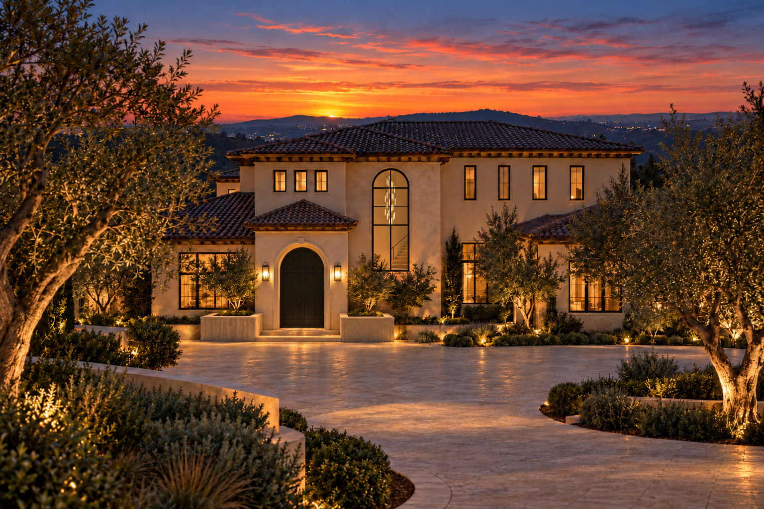 A Spanish Colonial house at sunset with clay tile roofs, a closed arched black metal courtyard gate between lanterns and two low rectangular stone planters, a tall arched stair window with a dense centered cascade of slender tube pendants about one-third of the window width, ending just past its middle above a visible stair flight, glowing windows, and uplit olive trees around a pale stone motor court.